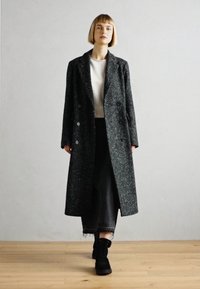 Dvouřadový vlněný kabát tmavě šedé barvy s texturovaným vzorem herringbone. Model nosí černé široké kalhoty a černé kotníkové boty.
