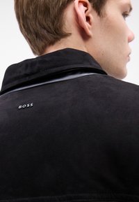 Giovane mostrato di spalle che indossa un giubbotto in suede nero con un colletto in pelle e il logo "BOSS" sulla schiena.