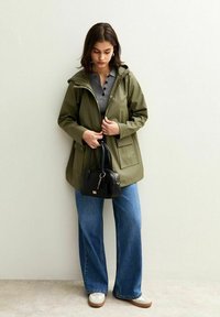 Chaqueta impermeable verde oliva con capucha, dos bolsillos frontales, combinada con una camiseta gris, jeans anchos azules y un pequeño bolso negro.