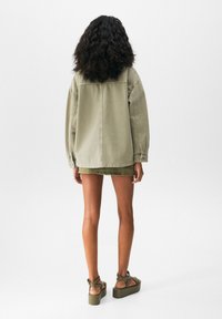 Mujer con cabello rizado de pie de espaldas, vistiendo una chaqueta beige oversized, una falda verde oliva y sandalias de plataforma, frente a un fondo liso.