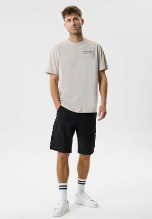 Homme debout les mains dans les poches, portant un t-shirt beige, un short cargo noir, des chaussettes blanches à rayures noires et des baskets blanches sur fond blanc.