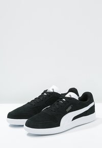 Schwarze Wildledersneaker mit einer weißen Gummisohle, versehen mit einem weißen Seitenstreifen und schwarzen Schnürsenkeln. Akzentuiert mit einem Logo auf der Zunge.