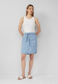 Lichtblauwe denim rok met een strikriem, rechthoekige vorm, subtiele stiksels en een zoom net boven de knieën. Witte, aansluitende tanktop gedragen.
