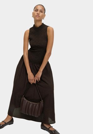 Top senza maniche nera con texture abbinato a una lunga gonna a quadri marrone. Tiene in mano una borsa a pieghe marrone. Scarpe flat completano l'outfit.