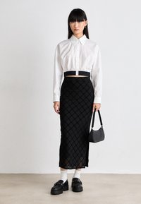 Chemise blanche à manches courtes et coupe courte associée à une jupe à motifs géométriques noirs. Des chaussures à plateforme noires et un petit sac noir complètent le look.