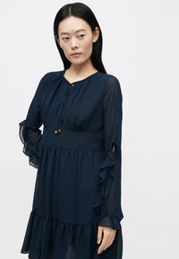 Μπλε-ναυτικό φόρεμα από σιφόν με βολάν στο τελείωμα και στις μανσέτες, με στρογγυλή λαιμόκοψη και σκοινάκι στο μπροστινό μέρος.