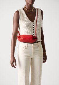 Rød skinncrossbody-veske med en myk, rund form, med folder og et tynn, justerbar skulderrem. Modellen har på seg en hvit strikket topp og beige jeans.