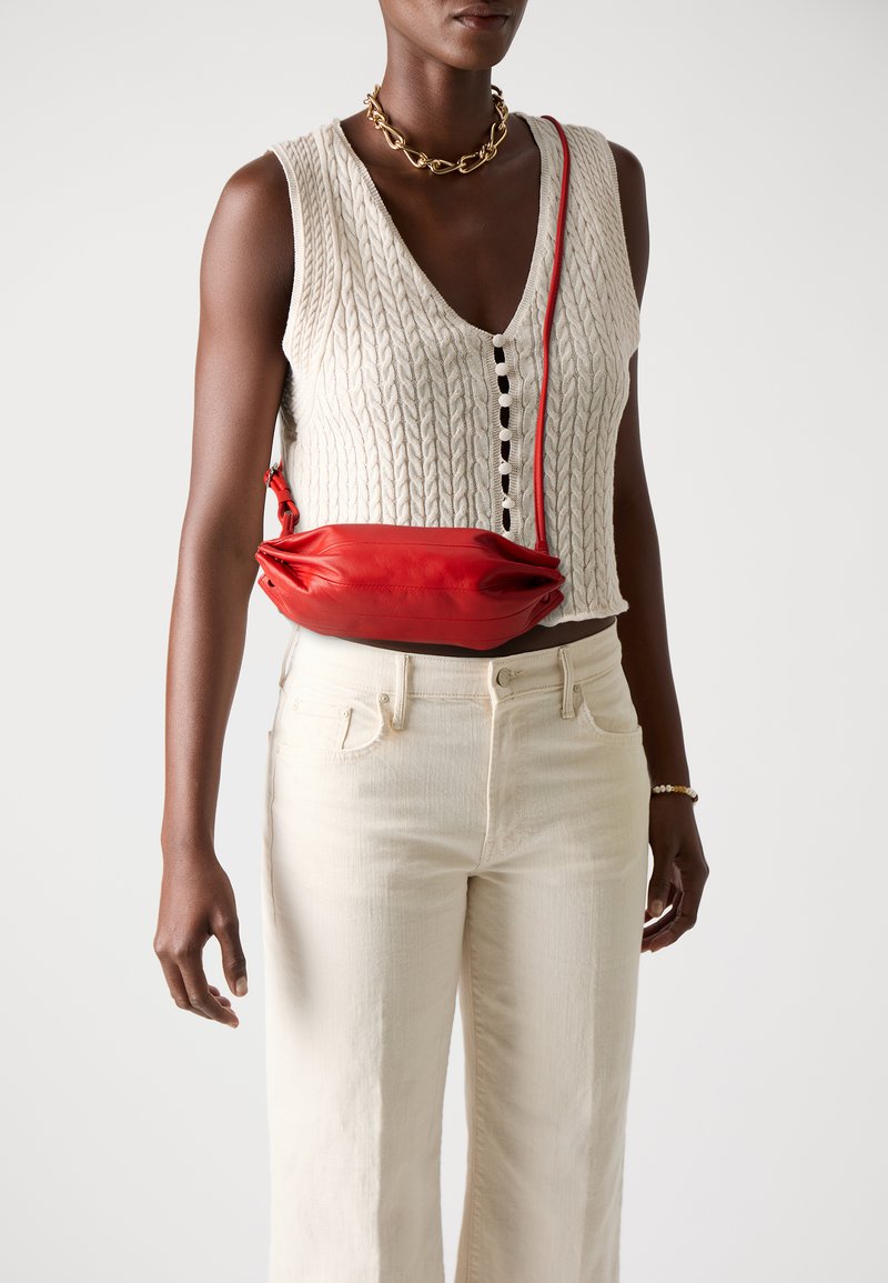 Rød skinncrossbody-veske med en myk, rund form, med folder og et tynn, justerbar skulderrem. Modellen har på seg en hvit strikket topp og beige jeans.