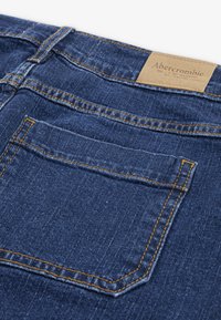 Denim kavbojke v temno modri barvi, s klasičnim krojem, oranžnim šivanjem, zadnjim žepom in svetlo rjavim usnjenim etiketom na pasu.