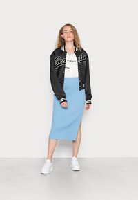 Casaco bomber de satin preto com riscas brancas, texto gráfico. T-shirt branca com padrão tie-dye, saia midi de malha azul claro com fenda lateral. Ténis brancos.