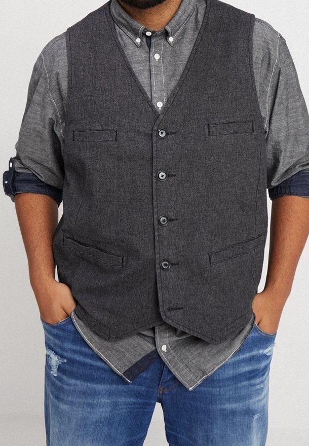 Homme portant un gilet boutonné gris foncé par-dessus une chemise grise à manches longues retroussées et un jean bleu usé, les mains dans les poches.