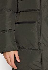 Ciemnozielona kurtka puchowa z fakturowanym wykończeniem, poziomym pikowanym wzorem oraz przednią kieszenią z czarnym wykończeniem.