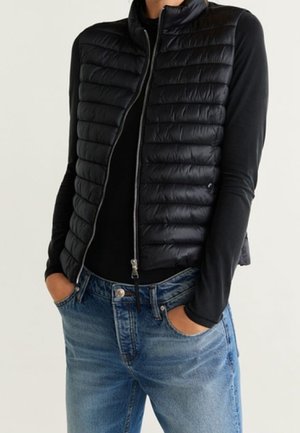 Veste sans manches matelassée noire portée sur un haut noir à manches longues, mains dans les poches d'un jean en denim bleu sur fond uni.