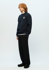 Navy crewneck sweatshirt with a logo, worn over a white shirt, paired with loose black trousers and black footwear.