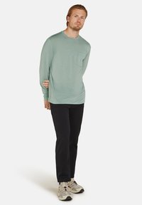 Hellgrünes Langarmshirt mit einer Brusttasche, kombiniert mit schwarzen Hosen und beigen Sportschuhen. Einfaches Design mit lockerer Passform.