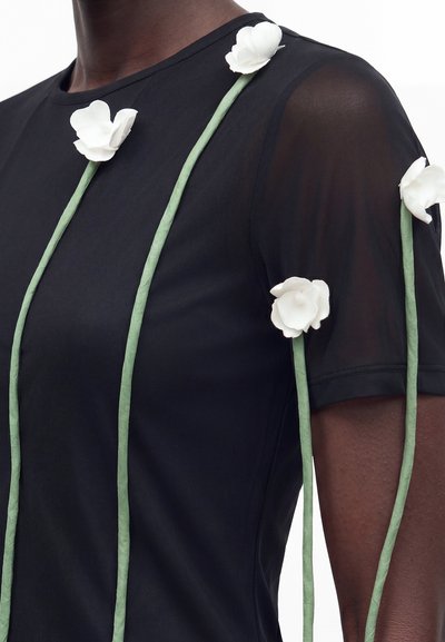 Μαύρο κοντομάνικο τοπ με διαφανή μανίκια, διακοσμημένο με μακριά πράσινα κλαδιά και λευκά υφασμάτινα λουλούδια προσαρτημένα κατά μήκος του μπροστινού μέρους και των μανικιών.