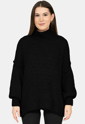 Mladá žena s dlouhými hnědými vlasy, oblečená v černém oversized roláku a malých zlatých kruhových náušnicích, stojící před jednoduchým pozadím.