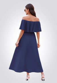 Abito maxi blu navy con spalle scoperte, dal corpetto svasato e cinturino elastico; texture liscia; abbinato a sandali bianchi con tacco alto e fascia.