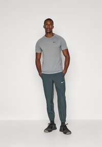 Tricou athletic gri cu mânecă scurtă și logo black, asortat cu pantaloni conici de culoare verde închis și pantofi sport negri. Material fin și design ajustat.