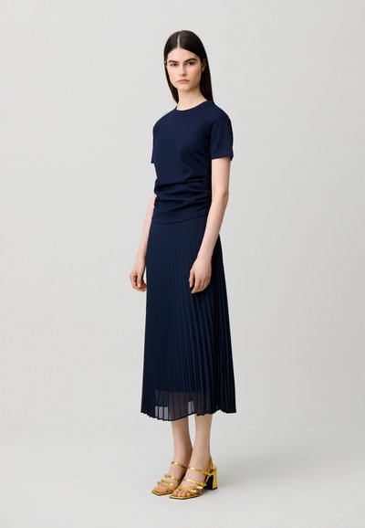 Donna con top blu navy a maniche corte e gonna midi plissettata con sandali dorati con tacco a blocchetto, in piedi davanti a uno sfondo semplice.
