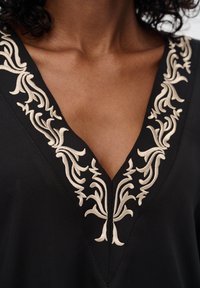 Schwarzes Kleidungsstück mit tiefem V-Ausschnitt, verziert mit kunstvoller, cremefarbener Blumenstickerei entlang der Kanten. Glatte Textur, lässiges Design.