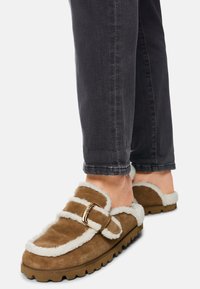 Braune Wildleder-Slipper mit weißem Kunstfellfutter, verstellbarem Riemen und goldfarbenen Beschlägen, kombiniert mit dunklen Jeans. Strukturierte Sohle sichtbar.