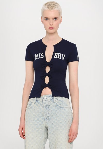 Navy short-sleeve shirt with a round neckline, keyhole opening, and bow accents. Features a printed logo and is paired with light blue patterned jeans.