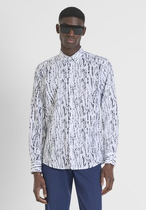 Witte longsleeve shirt met zwart abstract patroon, met knoopsluiting kraag en gestructureerde stof; gecombineerd met marineblauwe broek en zwarte zonnebril.