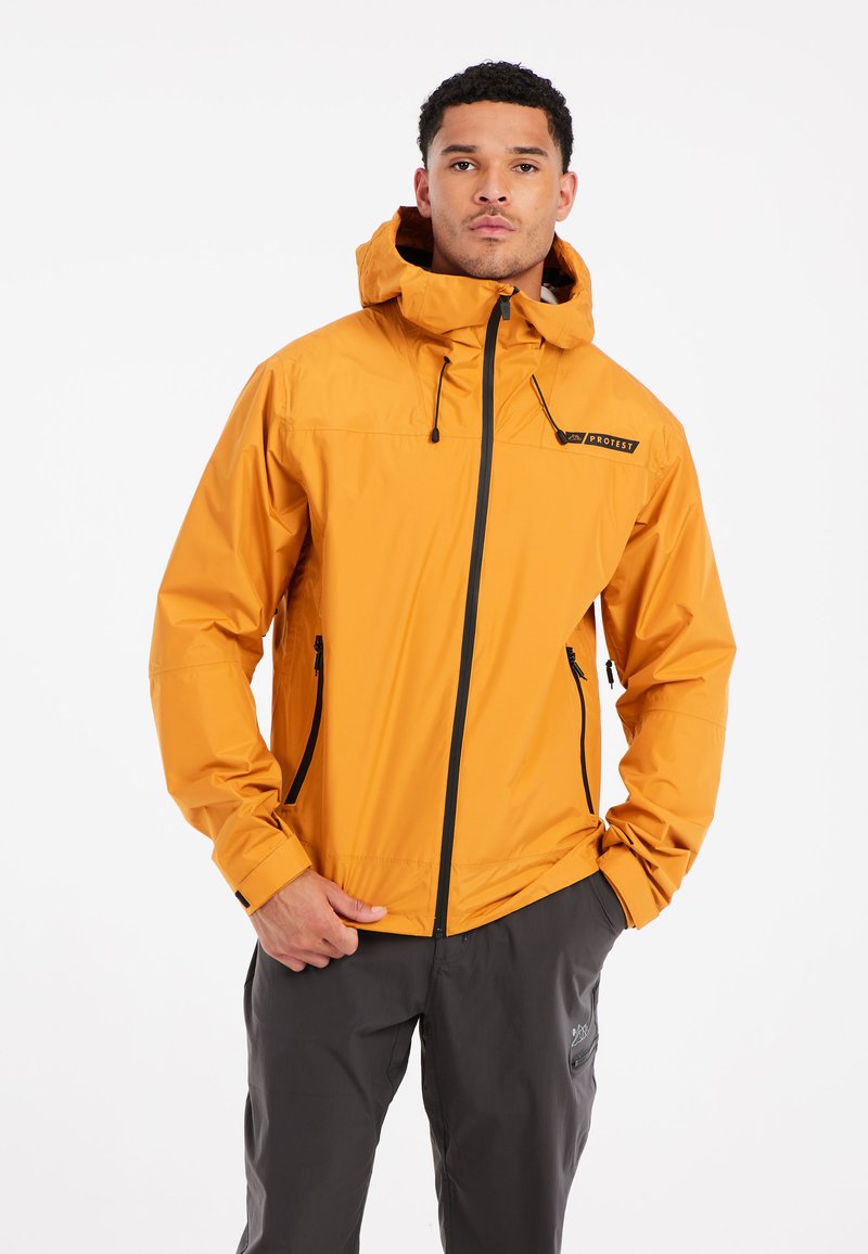 Man in een feloranje hoodie met zwarte ritsen en donkergrijze broek, staande met één hand in de zak tegen een witte achtergrond.