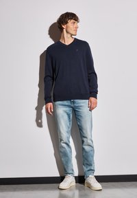 Marinblå, långärmad tröja med V-hals och ribbade ärmslut, tillsammans med ljusblå, urtvättade jeans och vita sneakers.