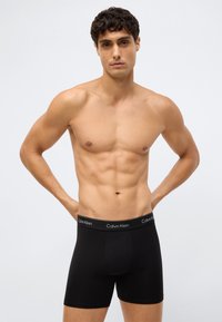 Atletische man staat tegen een effen achtergrond en draagt zwarte Calvin Klein boxershorts met zijn handen op zijn heupen.
