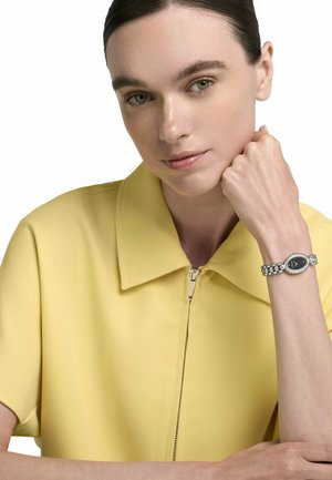 Mujer con una camisa amarilla de cuello, apoyando el mentón en su mano, mostrando un reloj de pulsera plateado con esfera ovalada y fondo negro.