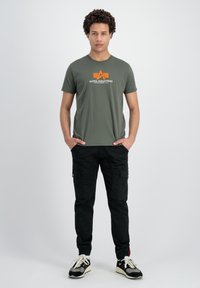 Olīvu zaļš T-krekls ar "ALPHA INDUSTRIES" logo un oranžu zvaigzni; melnas piedurkņu bikses ar sānu kabatām; melni sporta apavi ar baltām auklām.