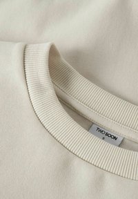 Beiger Baumwollpullover mit einem gerippten Rundhalsausschnitt, versehen mit einem Etikett "TWO SOON" und der Größe "S." Glatte Textur, minimalistisches Design.