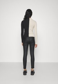 Kvinna med långt svart hår som bär en svart och beige vertikalt delad tröja, svarta åtsittande byxor och svarta platåskor, står med ryggen mot kameran.