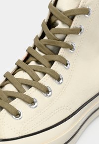 Zapatilla alta de lona color beige con cordones verdes olivo, ojales plateados, suela negra y sutiles detalles de costura.