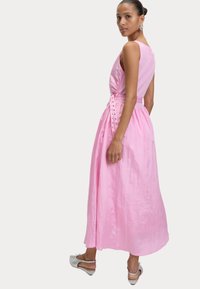 Robe maxi rose avec un corsage ajusté, tissu texturé et un accent de lien sur le côté. La jupe tombe jusqu'à la cheville avec une douce brillance. Chaussures argentées.