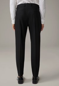 Pantaloni neri eleganti con una vestibilità su misura, dotati di due tasche posteriori, pinces e una texture liscia, abbinati a scarpe nere.
