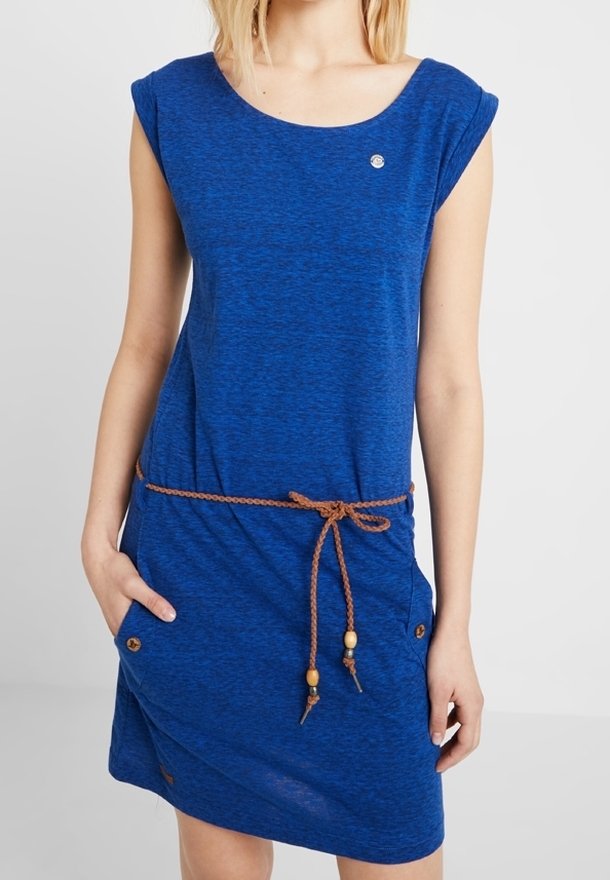 Femme portant une robe bleue jusqu'aux genoux avec des manches courtes et une fine ceinture tressée marron nouée à la taille, debout avec une main dans la poche.