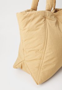 Borsa tote trapuntata in morbido tessuto beige. Presenta due manici robusti e una base triangolare spaziosa con dettagli di cucitura visibili.