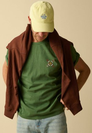 Man wearing light yellow cap with "LA DOLCE VITA," green t-shirt with small emblem, light jeans, and brown sweatshirt draped over shoulders.