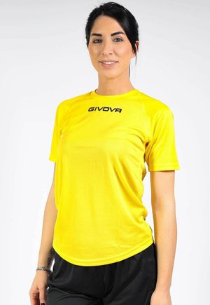 Mujer con el pelo oscuro recogido, vistiendo una camiseta deportiva Givova de color amarillo brillante y pantalones negros sobre un fondo blanco.