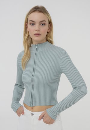 Top azzurro chiaro a costine con maniche lunghe, colletto alto e zip frontale. Design aderente e lunghezza cropped, con polsini a foro per il pollice.