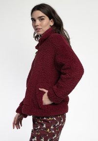 Chaqueta de forro polar burdeos con cuello, superficie texturizada, dos bolsillos frontales y mangas largas; llevada sobre pantalones de pijama estampados.