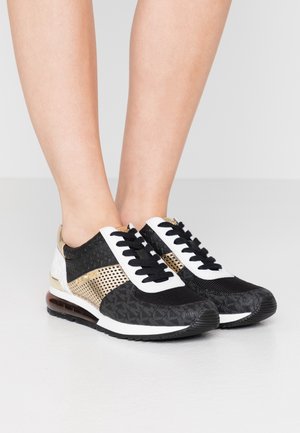 Czarno-biało-złote sneakersy sznurowane noszone na stopach, z wzorzystymi teksturami i widoczną podeszwą z poduszką powietrzną.