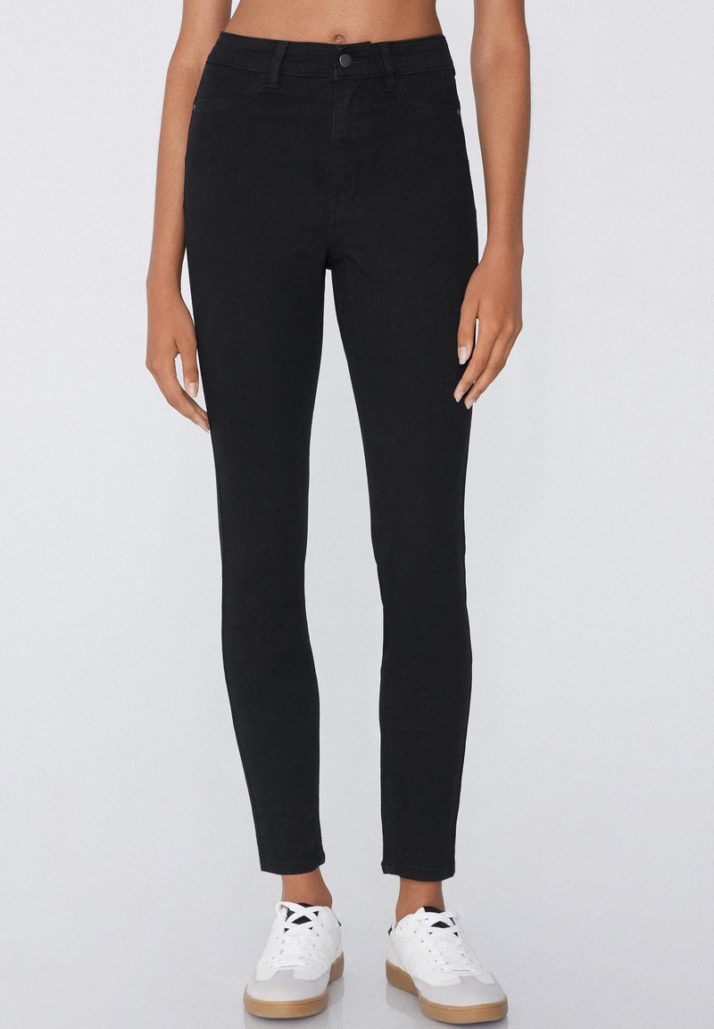 Jean skinny noir avec une taille mi-haute, doté d'une fermeture à bouton à l'avant et d'un design à cinq poches, associé à des baskets blanches.