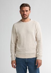 Crème-kleurige trui van textuurstof, met een ronde halslijn en ribgebreide manchetten en onderrand. Gedragen met blauwe denim jeans.