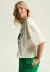 Luźna kremowa koszulka z krótkimi rękawami, z zielonym nadrukowanym tekstem i prostym krojem, zestawiona z zielonymi spodniami. Gładka faktura materiału.