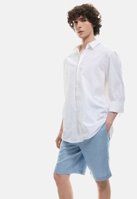Camisa blanca de botones con mangas enrolladas, combinada con pantalones cortos de color azul claro. La camisa tiene un cuello clásico y un bolsillo en la parte frontal.