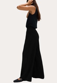 Pantalones de pierna ancha de canalé negros con cintura alta y tejido texturizado. Combinados con una blusa sin mangas negra y zapatos de punta, que presentan una hebilla decorativa.